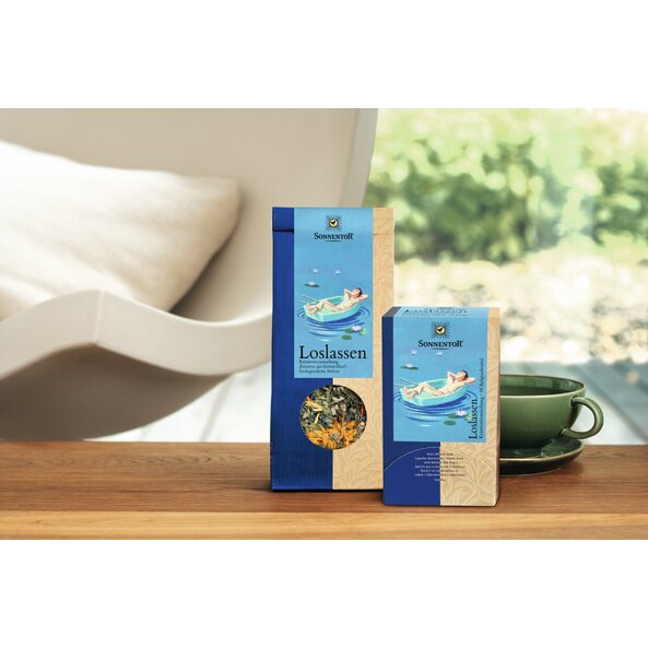 Foto von einer Packung Loslassen Tee auf einem Tisch, links davon steht eine Packung Loslassen Tee lose und rechts eine grüne Tasse.