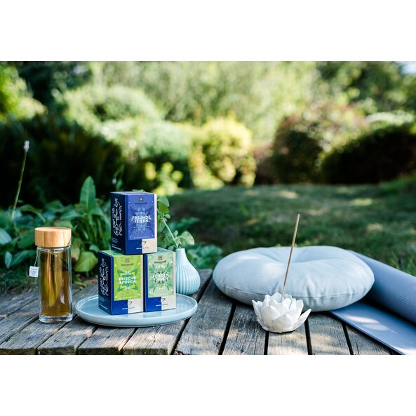 The picture shows three Happiness Is tea packs outside. Next to them is a bottle filled with tea. There is also a pillow and a yoga mat next to it.